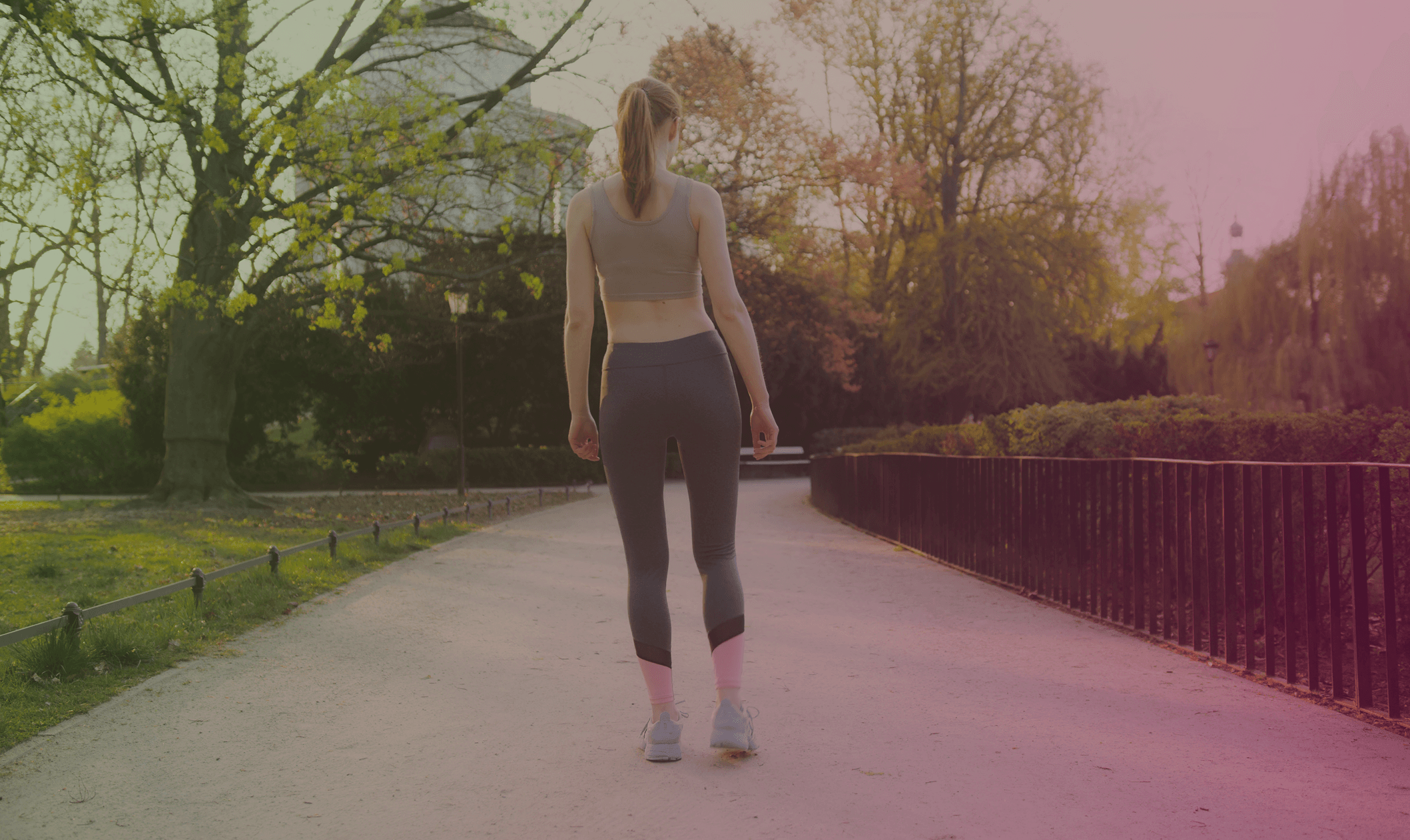 A woman in a sports bra and grey leggings with pink accents walks on a park path, her back to the viewer, under the warm afternoon light.